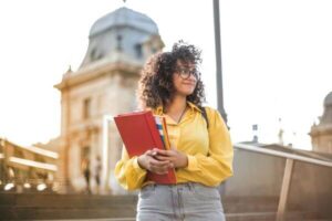 Young woman with a red folder and backpack smiling on a university campus in Spain, ready to study abroad.