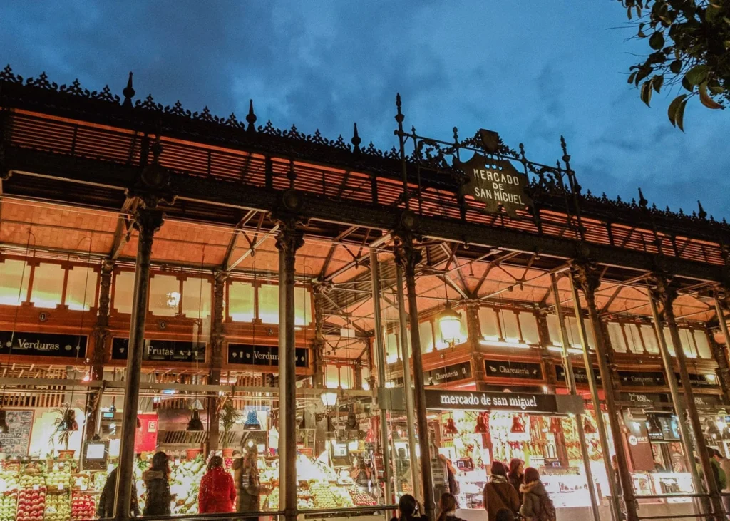 Night view of Mercado de San Miguel in Madrid, with glowing interior lights and people shopping for fresh food and tapas.