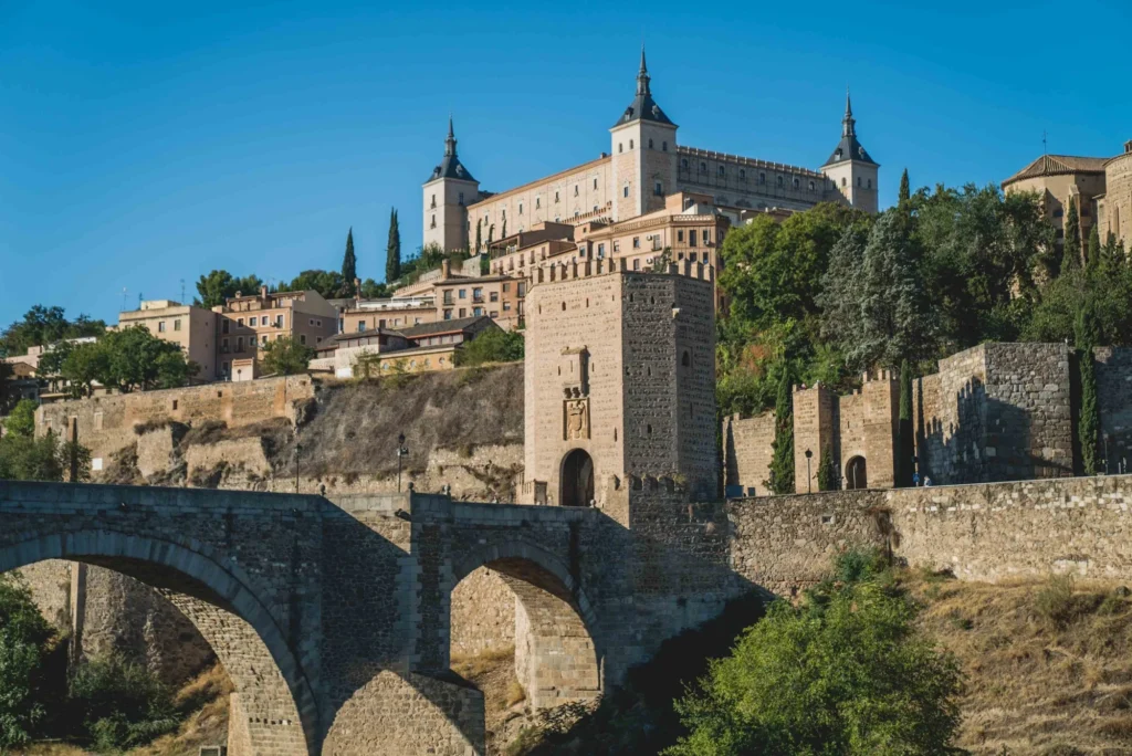 Historic view of the Alcázar and Puerta de Alcántara in Toledo, Spain, with medieval architecture and stone bridge.