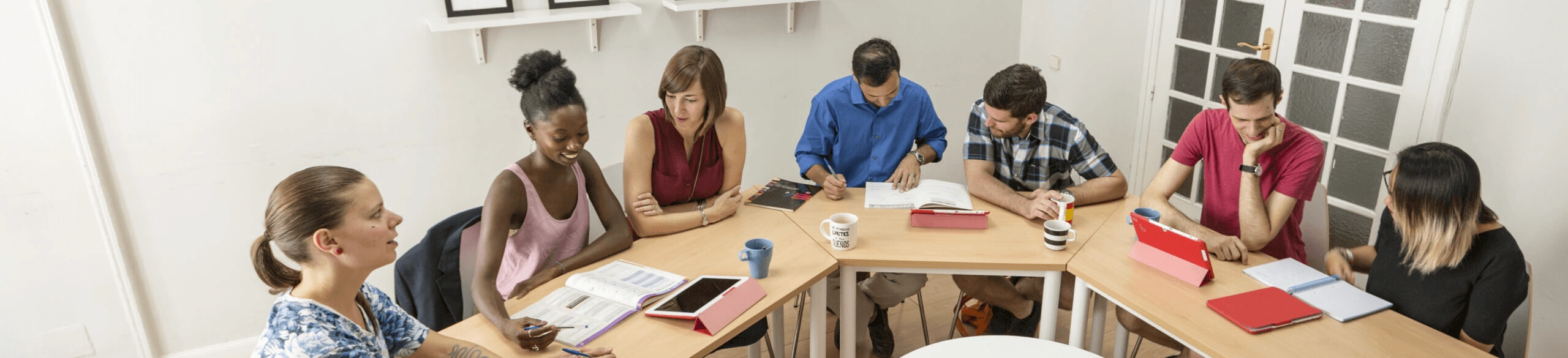 Diverse group of adult students engaged in a Spanish language class at LAE Madrid, seated in a collaborative classroom setting with study materials and tablets