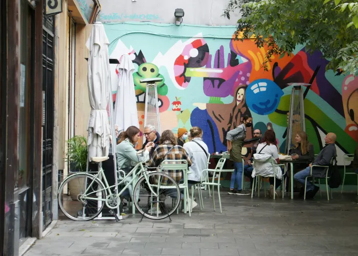 A group of people sitting at an outdoor terrace next to a bicycle, with a colorful artistic mural in the background on a Madrid street.