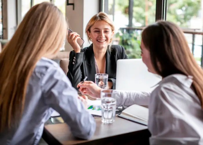 Three professional women sitting at a table in a bright office or cafe setting, smiling and discussing work with a laptop and notebooks open.