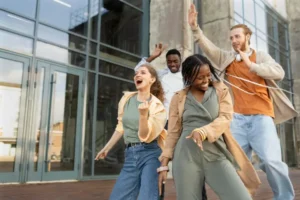A joyful group of four young diverse students dancing and laughing outside a modern glass building in Madrid, celebrating a breakthrough.