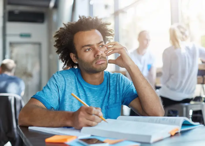 A young man with curly hair wearing a blue t-shirt, sitting at a desk and looking thoughtful while writing in a notebook with a pencil.