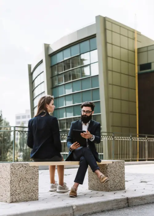 A professional woman and a man with a beard and glasses sitting on an outdoor stone bench, reviewing information on a tablet in front of a modern glass building.