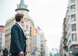 : A young professional man in a suit standing on a Madrid street, holding a coffee cup and looking at the classical European buildings around him.