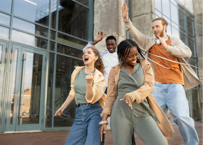 A joyful group of four young diverse students dancing and laughing outside a modern glass building in Madrid, celebrating a breakthrough.