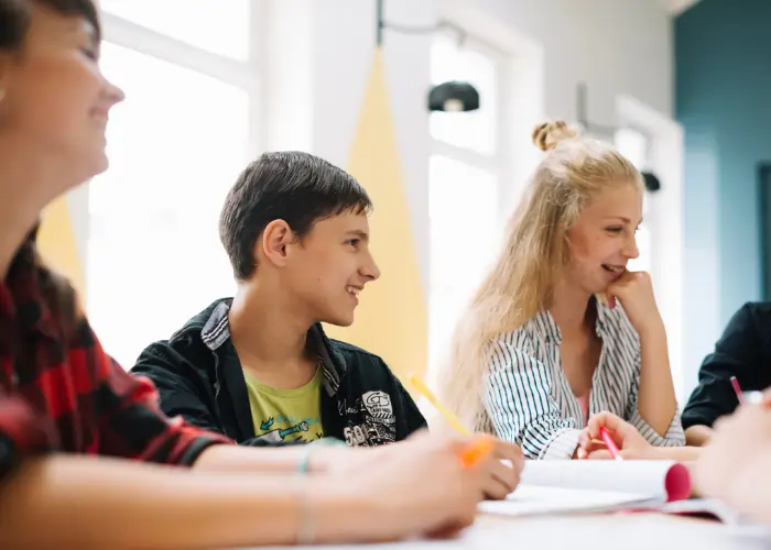 A small group of three diverse students in a bright BLC Spain classroom, smiling and engaged in a collaborative discussion while taking notes.