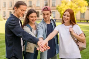 A diverse group of four young Master's students standing in a circle outdoors on a campus lawn, smiling and joining their hands together in the center.