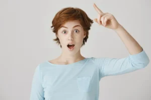 A young woman with short red hair and freckles looking surprised and inspired, pointing her finger up as if she just had a breakthrough idea.