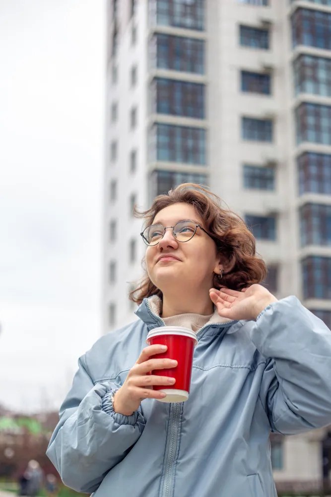 young professional with glasses holding a red coffee cup against a modern Madrid building, illustrating the city's anti-burnout culture.