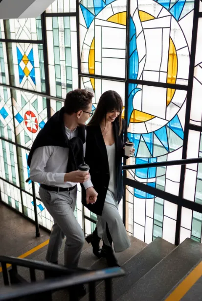Two young professionals talking and walking up stairs past a colorful stained-glass window at BLC Spain in Madrid.