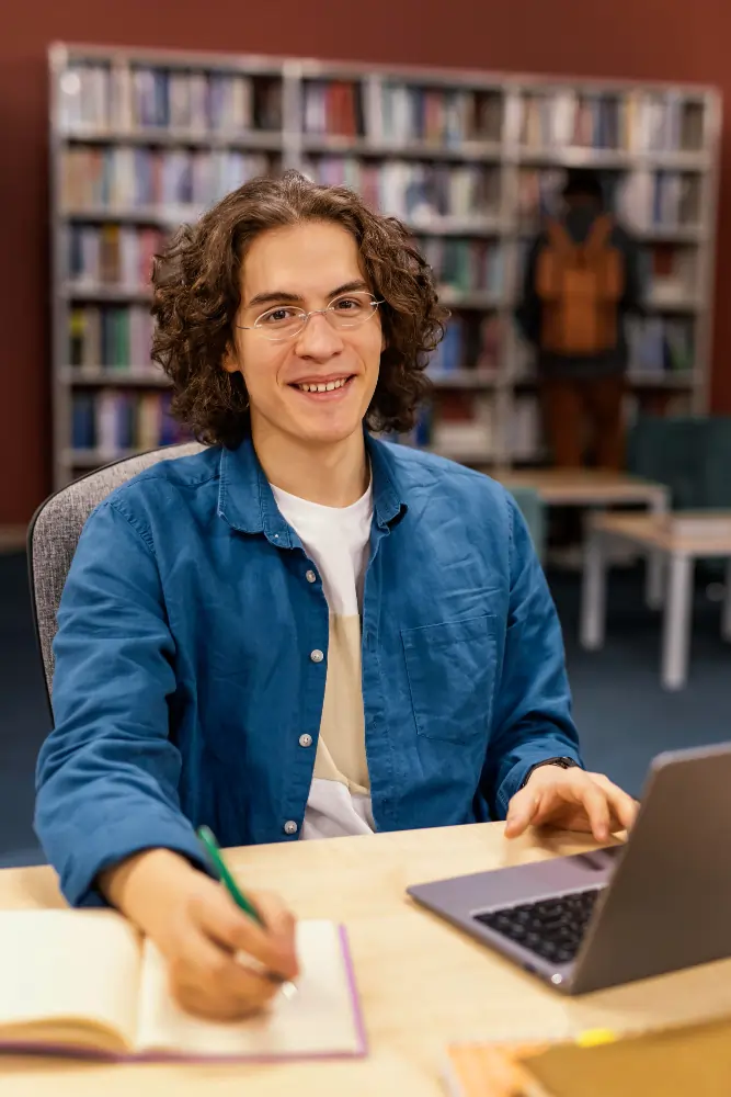 A smiling student sits with a laptop and notebook in a modern Spanish university library, illustrating the transition to higher studies for residency in Madrid.