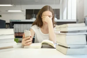 A stressed young woman in a library covering her face while looking at her phone, representing an English language assistant receiving news of the 2026 program cancellation.