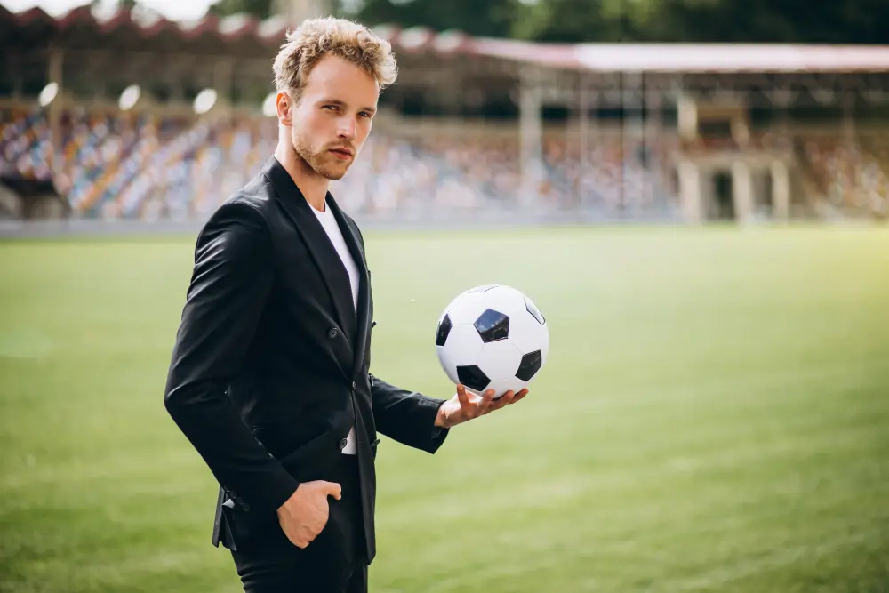 A blonde businessman in a sharp black suit holding a classic black-and-white football in his hand while standing alone on an empty green stadium pitch.