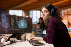 A focused woman wearing headphones working at a dual-monitor computer workstation displaying complex neural network and AI code diagnostics.