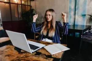 A young woman with glasses celebrating excitedly with her arms raised in front of an open laptop at a cafe table with a notebook.