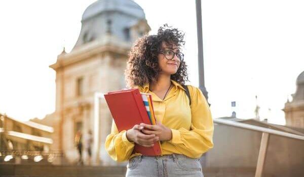 Young woman with a red folder and backpack smiling on a university campus in Spain, ready to study abroad.