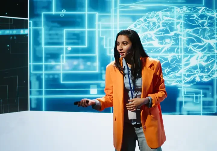 professional woman in an orange blazer giving a presentation on artificial intelligence with a large digital brain graphic in the background during a conference in Madrid.