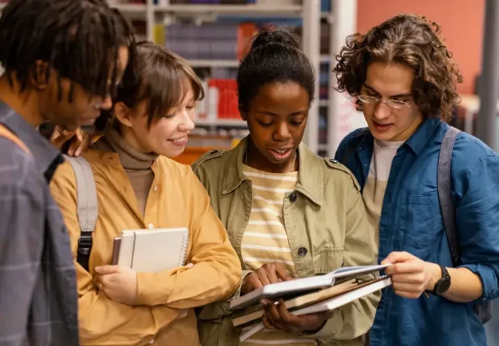A diverse group of four young professionals standing together in a library, looking at a book and discussing their studies.