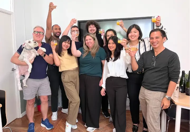 : A diverse group of smiling professionals and a dog raising glasses for a toast in a modern office setting at BLC Spain in Madrid.