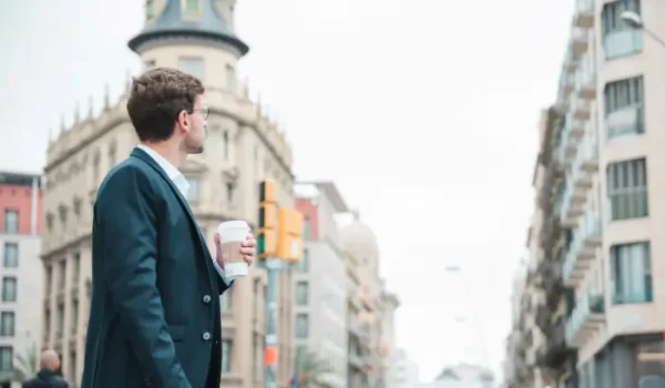 : A young professional man in a suit standing on a Madrid street, holding a coffee cup and looking at the classical European buildings around him.