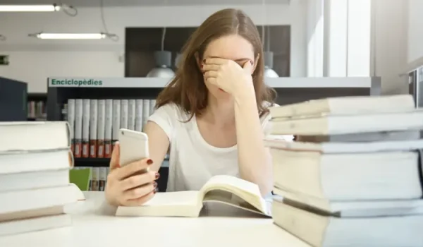 A stressed young woman in a library covering her face while looking at her phone, representing an English language assistant receiving news of the 2026 program cancellation.