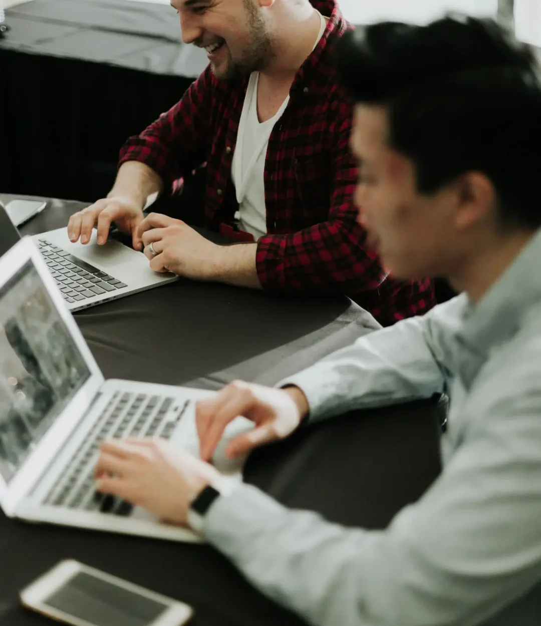 Two men working on laptops at a dark table; one is smiling and wearing a plaid shirt, the other is typing in a light blue shirt.