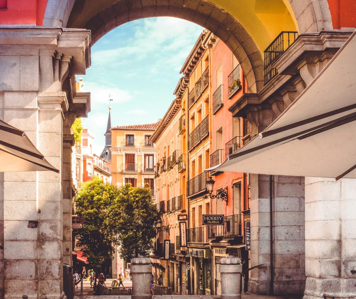 Charming view through an archway in Plaza Mayor, Madrid, showing colourful buildings, local shops, and street life.